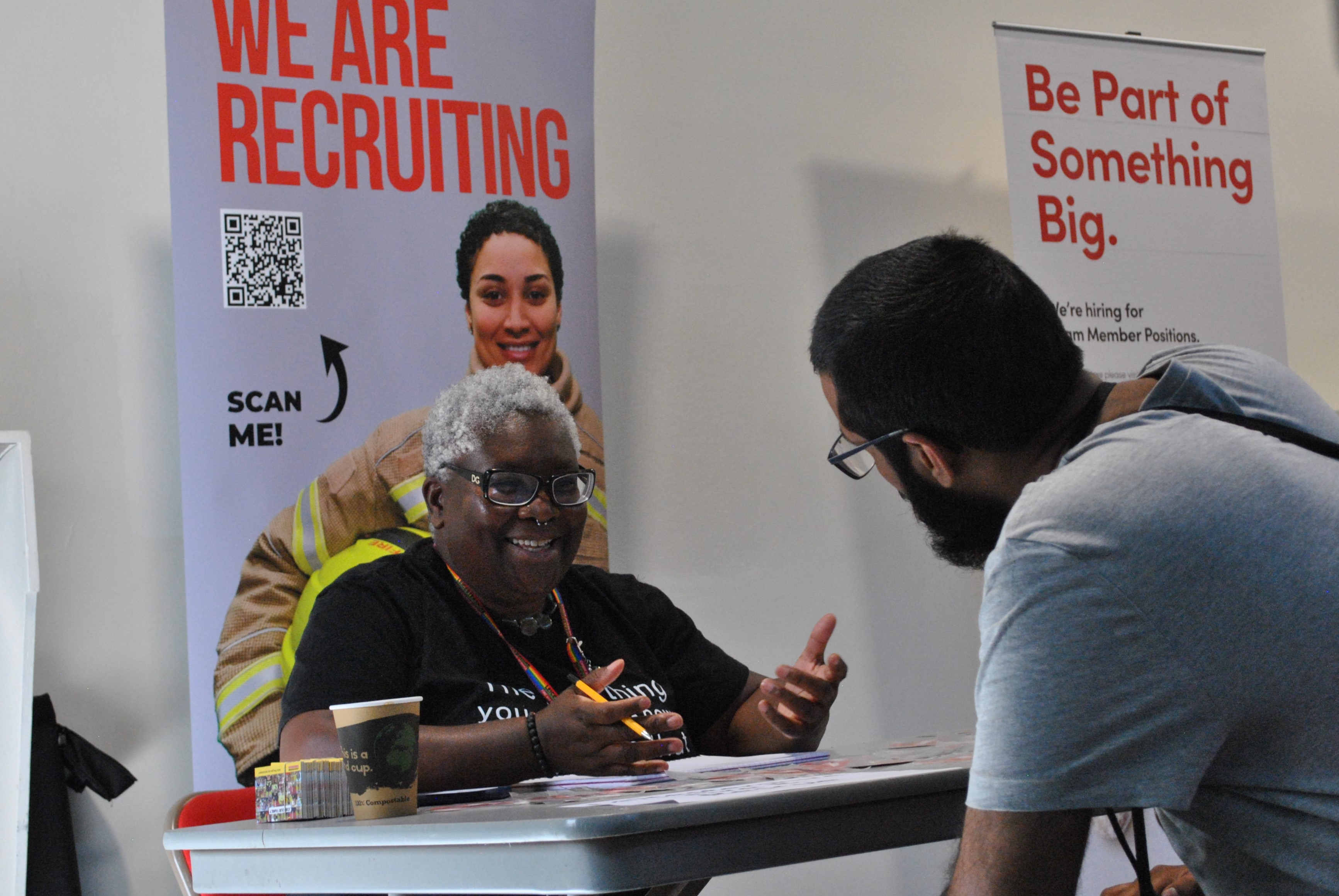 A woman interviews a man with a recruitment poster behind her