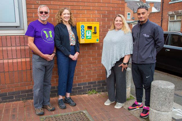 Two men and two women standing by a defibrillator on an outside wall.
