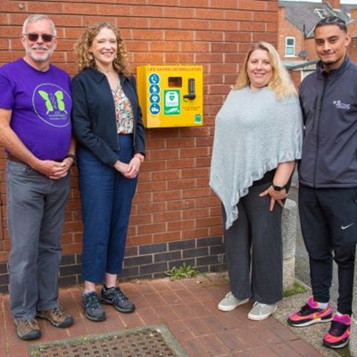 Two men and two women standing by a defibrillator on an outside wall.