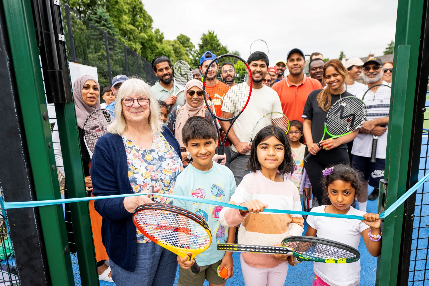 A woman cuts a blue ribbon to open a tennis court with children next to her.