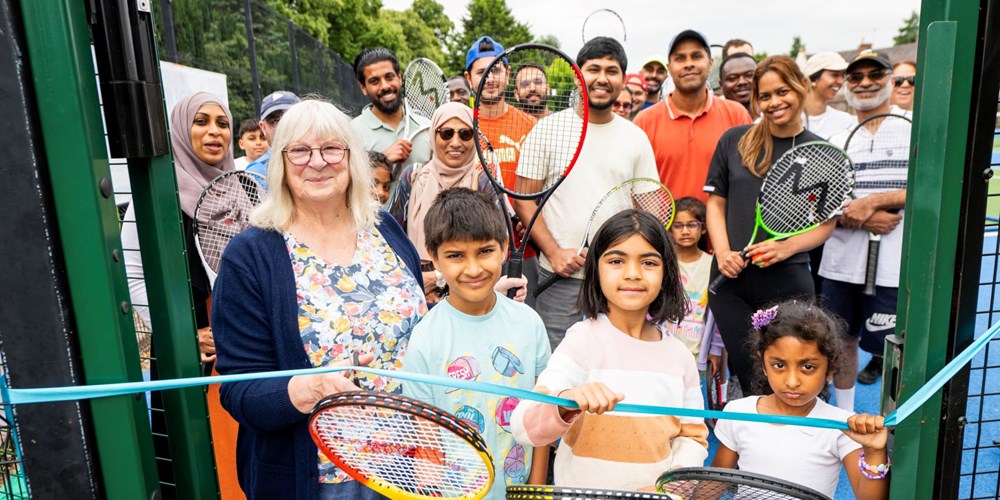 A woman cuts a blue ribbon to open a tennis court with children next to her.