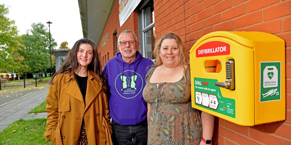 A man in a purple sweatshirt stands between two women next to a defibrillator on the external brick wall of a building