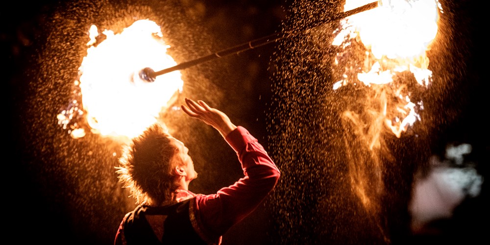 A man holds a stick above his head with flames at each end, against the backdrop of a night sky.