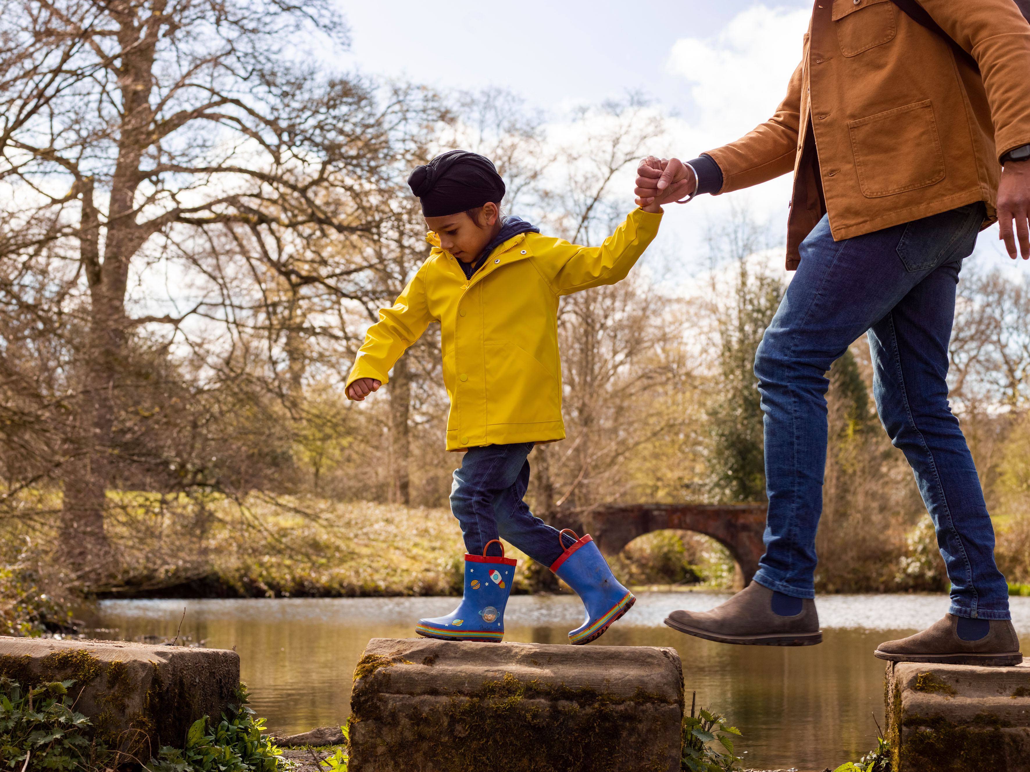 A young boy wearing a turban holds the hand of an adult as they walk across stepping stones on water.