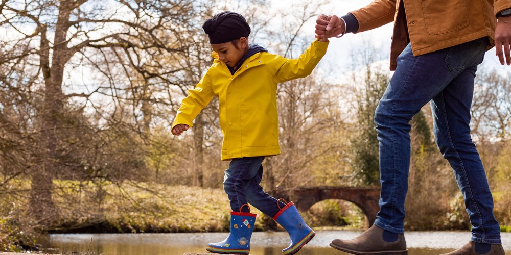A young boy wearing a turban holds the hand of an adult as they walk across stepping stones on water.