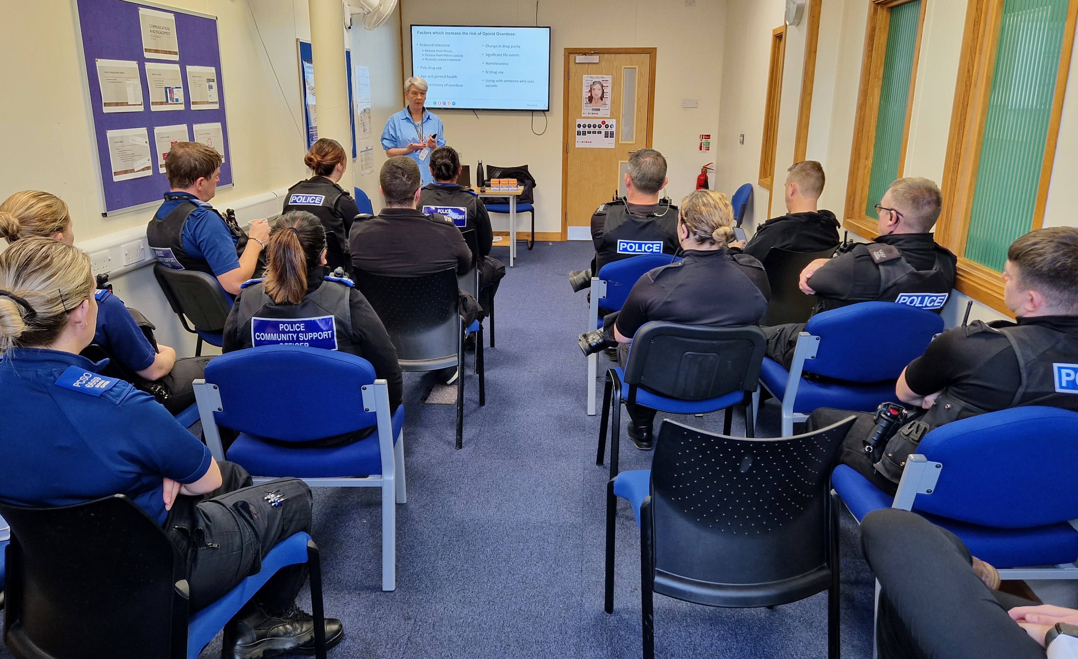 Police officers sit in rows of chairs with their back to the camera watching a training instructor at the front of the room.