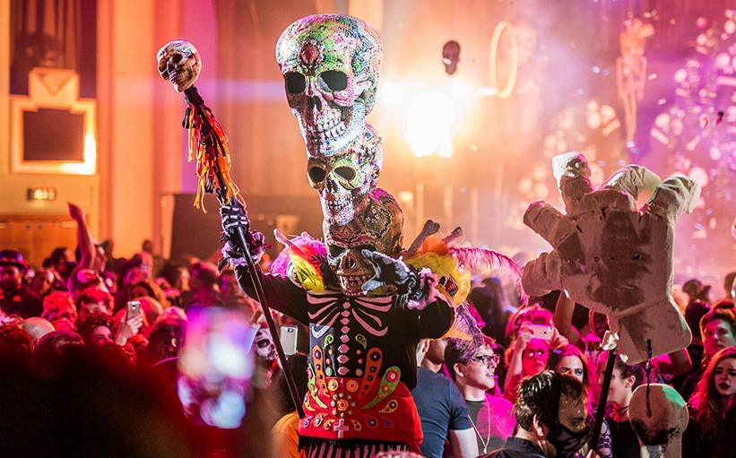 A spooky character in costume with large skulls perched on his head entertains crowds.
