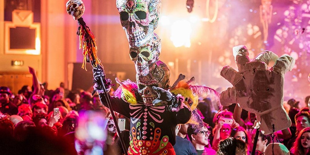A spooky character in costume with large skulls perched on his head entertains crowds.