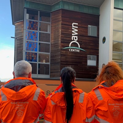 Three people wearing orange outreach jackets have their back to the camera, with the Dawn Centre hostel in the background.