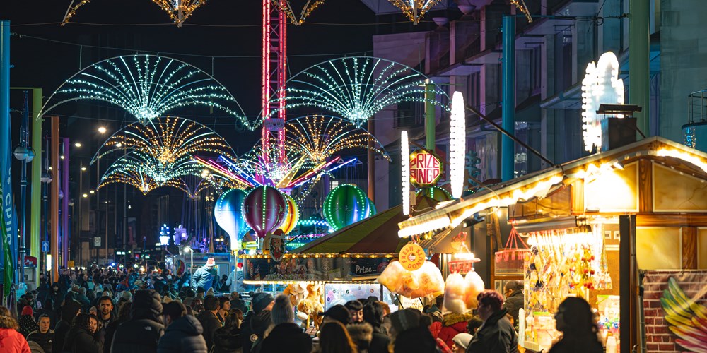 Brightly coloured Christmas lights stretch across a street with wooden market stalls in the foreground