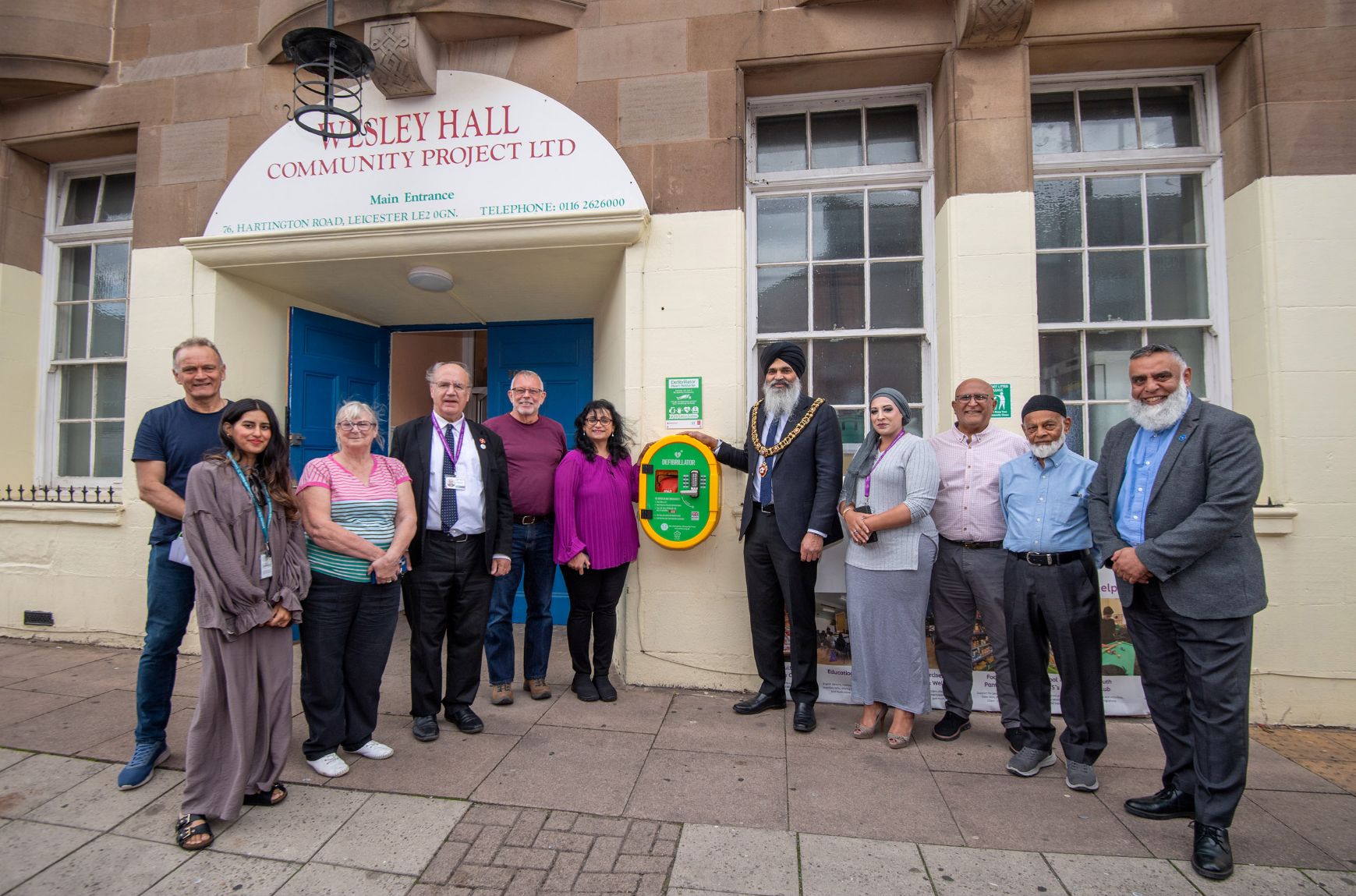 A group of people stand either side of a defibrillator machine fitted to an external wall on a building.
